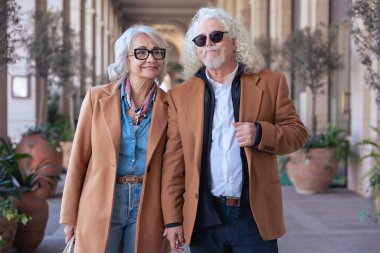 Güneş gözlüğü ve şık ceketler takan gülümseyen yaşlı bir çiftin dikey portresi. Şehir ortamında zarafet, mutluluk ve bağlantı göstererek güvenle dışarı çıkarlar.. 