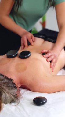 Sıcak taş masajının yakın görüntüsü, rahatlatıcı bir terapi seansı sırasında sırtüstü uzanmış siyah taşlar. Spa, sağlık, iyileşme ve alternatif sağlık konseptleri için mükemmel. Dikey