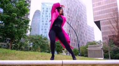 Pembe palto giyen kıvırcık saçlı Afrikalı Amerikalı dansçı şehir ortamında hareketlerini ve enerjilerini ifade ediyor. Modern moda, modern dans ve sanatsal performans.