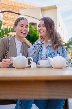 Dikey Portre İki genç beyaz kadın dışarıda çay içerken kameraya bakıp gülümsüyor. Güneşli bir günde rahat ve neşeli bir arkadaşlık, rahatlık ve sohbet anı..