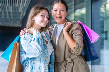 Renkli alışveriş torbaları taşıyan ve alışveriş merkezinde başarılı bir günün ardından gülümseyen iki beyaz kadın. Modern bir binanın dışında duruyorlar, birlikte eğleniyorlar stil ve neşe içinde. Dikey
