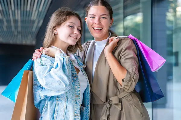 Renkli alışveriş torbaları taşıyan ve alışveriş merkezinde başarılı bir günün ardından gülümseyen iki beyaz kadın. Modern bir binanın dışında duruyorlar, birlikte eğleniyorlar stil ve neşe içinde. Dikey