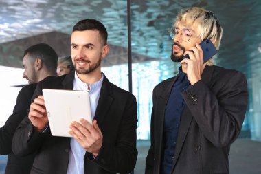 Modern bir ofiste iş birliği yapan takım elbiseli iki genç iş adamı. Biri telefonda konuşurken diğeri tablet kullanıyor, yansıtıcı camlar ve şık profesyonel bir atmosfer ile çevrili.. 