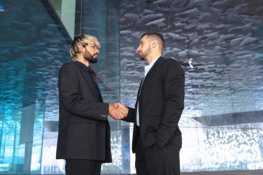 Modern cam bir ofiste takım elbiseli iki profesyonel işadamı el sıkışıyor. İş anlaşması, anlaşma ya da çağdaş tasarım ile kurumsal bir iç mekanda ortaklık. 