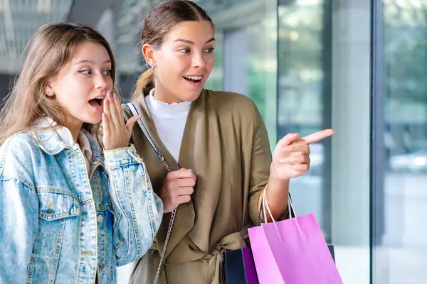 Alışveriş torbaları tutan ve vitrinlere bakan iki mutlu beyaz kadın. Şehirde alışveriş ve arkadaşlığın olduğu bir günde samimi ve neşeli bir şehir anı. 