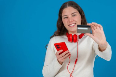 Beyaz kazaklı ve kırmızı kulaklıklı mutlu genç bir kadın kameraya bir kredi kartı gösterirken gülümsüyor ve elinde kırmızı bir akıllı telefon tutuyor, mavi arka planda, online alışverişi sembolize ediyor. Boşluğu kopyala
