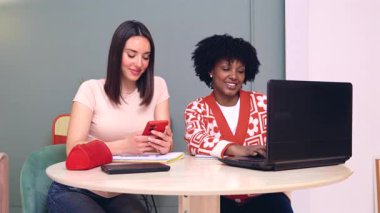 Birlikte çalışırken bilgisayar ve akıllı telefon kullanarak masada oturan gülümseyen öğrenciler. Samimi ve dost canlısı bir öğrenme alanında bir video çağrısına ya da çevrimiçi aktiviteye katılıyorlar.. 