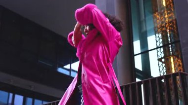 Pembe palto giyen kıvırcık saçlı Afrikalı Amerikalı dansçı şehir ortamında hareketlerini ve enerjilerini ifade ediyor. Modern moda, modern dans ve sanatsal performans.