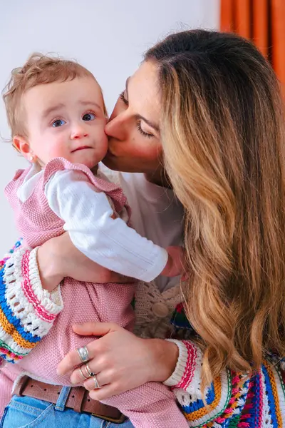 Sevecen annenin dikey portresi pembe tulumlu sevimli bebeğini öpmesi şefkatli aile sevgisi, bakıcılık bağı ve rahat bir ev içinde ebeveynlik hayatı göstermesi. 