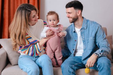 Kanepede oturup gülümseyen ve rahat bir anın tadını çıkaran genç ebeveynler. Sıcak aile sahnesi neşe, sevgi ve aile bağlarıyla dolu.. 