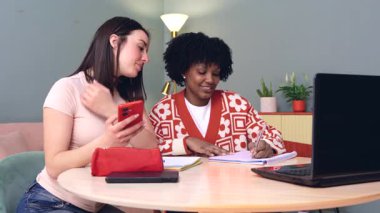Birlikte çalışırken bilgisayar ve akıllı telefon kullanarak masada oturan gülümseyen öğrenciler. Samimi ve dost canlısı bir öğrenme alanında bir video çağrısına ya da çevrimiçi aktiviteye katılıyorlar.. 