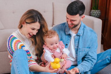 Oturma odasındaki kanepede gülümseyen ebeveynler pembe tulumlu küçük kızlarını sarı ördek ve yeşil kaplumbağa oyuncaklarıyla eğlendiriyorlar. Oyuncak aile bağlarını ve erken çocukluk gelişimlerini kapalı alanlarda yakalıyorlar.. 