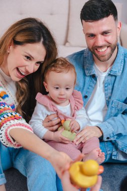 Oturma odasındaki kanepede gülümseyen ebeveynler pembe tulumlu kızlarını sarı ördekle eğlendirip neşeli aile bağlarını ve erken çocukluk gelişimlerini kapalı alanlarda yakalıyorlar. Dikey
