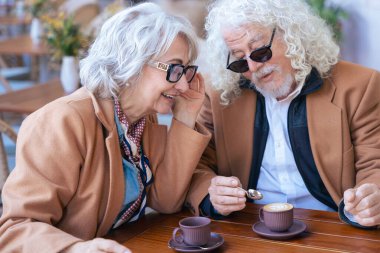 Gözlüklü ve kahverengi paltolu yaşlı bir çift açık hava kafesinde kahvenin tadını çıkarıyor. Sıcak bir anı paylaşıyorlar, gülümsüyorlar ve gündüzleri huzurlu, rahat bir atmosferde rahatlıyorlar..