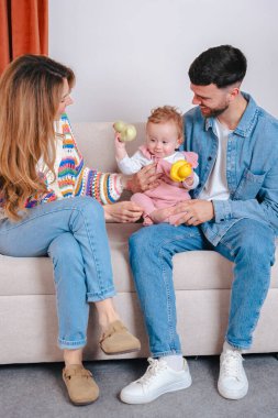 Kanepede gülümseyen ebeveynler pembe tulumlu, sarı ördek ve yeşil kaplumbağa oyuncaklarıyla küçük kızlarını eğlendiriyorlar. Oyuncak aile bağlarını ve erken çocukluk gelişimlerini içeride yakalıyorlar. Dikey