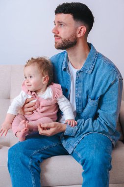 Kot gömlekli genç baba koltuğunda otururken bebeğini nazikçe tutuyor. Bebek, evde sakin ve samimi bir ebeveynlik anını yakalamak için tuhaf bir şekilde uzağa bakıyor.. 