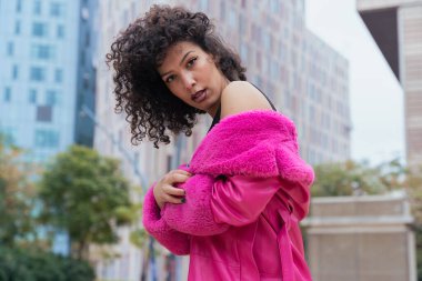 Kıvırcık saçlı, pembe ceketli, şehir ortamında hareket ve enerji ifade eden Afrikalı Amerikalı dansçı. Modern moda, çağdaş dans ve sanatsal performans.