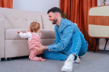 Kot pantolonlu genç bir baba, kanepe başında durup, destekleyici ebeveynlik, erken motor geliştirme ve evdeki rahat aile hayatını yakalama alıştırması yaparken pembe tulumlu bebeğine destek oluyor.. 