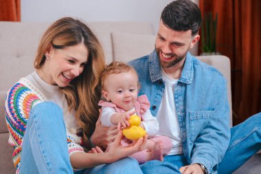 Oturma odasındaki kanepede gülümseyen ebeveynler pembe tulumlu küçük kızlarını sarı ördek ve yeşil kaplumbağa oyuncaklarıyla eğlendiriyorlar. Oyuncak aile bağlarını ve erken çocukluk gelişimlerini kapalı alanlarda yakalıyorlar.. 