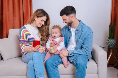 Anne, babasının kucağında oturan pembe tulumlu bebeğe akıllı telefon ekranı gösteriyor. Mutlu aile bağlarını, teknoloji kullanımını, erken öğrenme ve sıcak evde geçirilen aile sevgisini yakalıyor. 