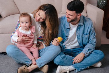 Genç ebeveynler sıcak, sıcak bir ev ortamında küçük kızlarıyla halının üzerinde oturuyorlar. Doğal etkileşim ve neşeli ruh hali aile, ebeveynlik ve sevgi temalı görsel içerik için mükemmeldir.. 