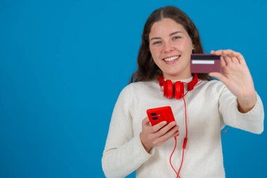 Beyaz kazaklı ve kırmızı kulaklıklı mutlu genç bir kadın kameraya bir kredi kartı gösterirken gülümsüyor ve elinde kırmızı bir akıllı telefon tutuyor, mavi arka planda, online alışverişi sembolize ediyor. Boşluğu kopyala