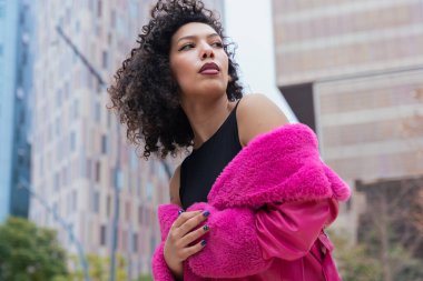 Kıvırcık saçlı, pembe ceketli, şehir ortamında hareket ve enerji ifade eden Afrikalı Amerikalı dansçı. Modern moda, çağdaş dans ve sanatsal performans.