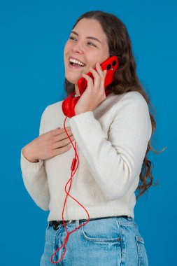 Kırmızı bir akıllı telefondan konuşan neşeli genç bir kadının dikey görüntüsü. Elini göğsüne koyup gülüyor. Parlak mavi bir arka plana yaslanıyor. Sıradan bir anda.. 