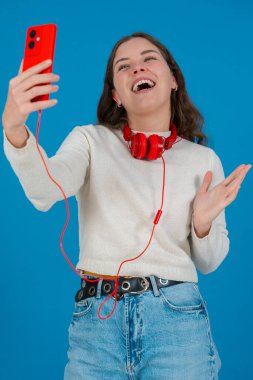 Kırmızı akıllı telefon ve kulaklıkla selfie çeken mutlu genç bir kadının dikey görüntüsü. Mavi bir arkaplan önünde, gündelik ve modern bir dijital anın tadını çıkararak güvenle gülümsüyor. Dikey
