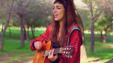 Uzun saçlı genç bir kadın güneşli bir parkta akustik gitar çalıyor, bir ağacın altında huzur içinde oturuyor. Doğal ortam, rahat atmosfer, müzik ve boş zaman atmosferi.
