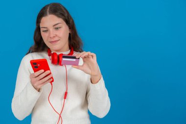 Beyaz kazaklı ve kırmızı kulaklıklı mutlu genç bir kadın kameraya bir kredi kartı gösterirken gülümsüyor ve elinde kırmızı bir akıllı telefon tutuyor, mavi arka planda, online alışverişi sembolize ediyor. Boşluğu kopyala