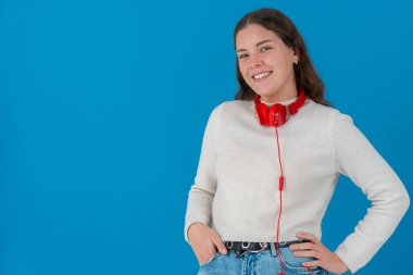 Beyaz kazak ve kot pantolon giyen, kırmızı kulaklık takan, gülümseyen ve elleri kalçasında poz veren, mavi arka planda duran, fotokopi çeken, kendine güvenen genç bir kadın.. 