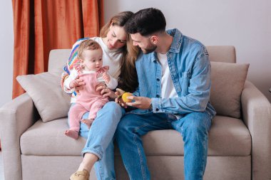 Anne babalar kanepeye yakın otururken bebekleri oyuncak tutarken şefkatli bir an. Sıcak ve rahat ev ortamları ebeveynlik, bağlılık, çocukluk ve duygusal bağ gibi konular için idealdir.. 