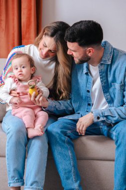 Anne babalar kanepeye yakın otururken bebekleri oyuncak tutarken şefkatli bir an. Sıcak ve rahat ev ortamları, ebeveynlik, bağlanma, çocukluk ve duygusal bağ gibi konular için idealdir. Dikey