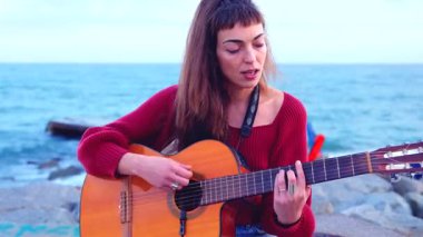 Akustik gitar çalan ve deniz kenarında şarkı söyleyen sanatçı genç bir kadın. Doğayla ve yumuşak doğal ışıkla çevrili sakin bir sahil ortamında duygusal bir performans sergiliyor.. 