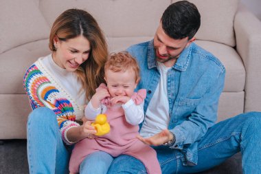 Oturma odasındaki kanepede gülümseyen ebeveynler pembe tulumlu küçük kızlarını sarı ördek ve yeşil kaplumbağa oyuncaklarıyla eğlendiriyorlar. Oyuncak aile bağlarını ve erken çocukluk gelişimlerini kapalı alanlarda yakalıyorlar.. 