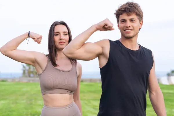 Gülümseyen genç adam ve kadın atletizm kıyafetleri içinde çimenli bir sahil parkında yan yana duruyor, kaslarını kaslarını kasıyor, sağlıklı, güçlü, ortaklık ve aktif yaşam tarzını sembolize ediyor.. 
