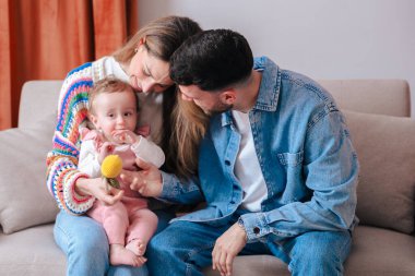 Anne babalar kanepeye yakın otururken bebekleri oyuncak tutarken şefkatli bir an. Sıcak ve rahat ev ortamları ebeveynlik, bağlılık, çocukluk ve duygusal bağ gibi konular için idealdir.. 