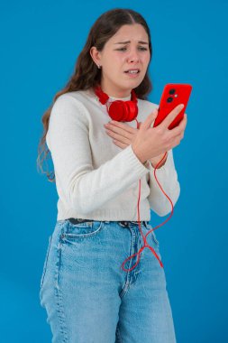 Beyaz kazaklı, elinde kulaklıkla kırmızı akıllı telefon tutan endişeli genç bir kadın, ekranı, alnı mavi arka planı izlerken şok ve stres içinde tepki veriyor.