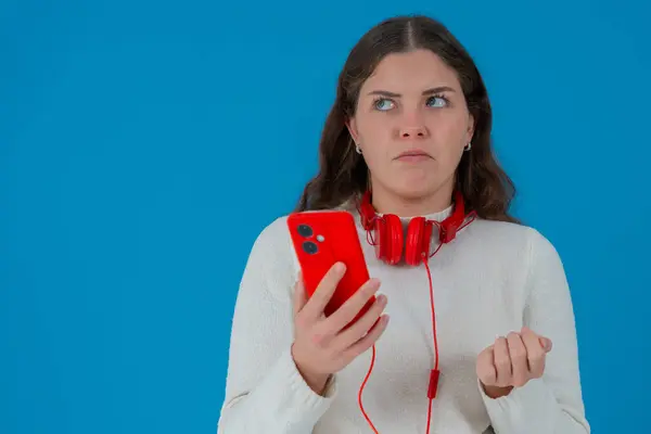 Beyaz kazaklı, kırmızı akıllı telefon ve kulaklık tutan endişeli genç bir kadın. Mavi arka planda izole bir şekilde ekrana bakarken olumsuz tepki veriyor. Boşluğu kopyala