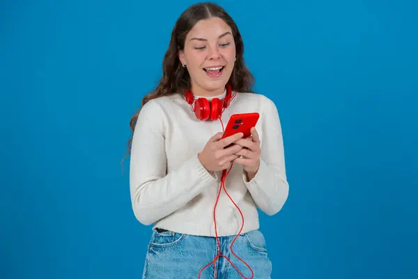 Boynuna kırmızı kulaklık takmış, akıllı telefonuna bakıp gülümseyen mutlu beyaz bir kadın. Mavi arka plan onun neşeli ve meşgul yüz ifadesini vurguluyor.. 