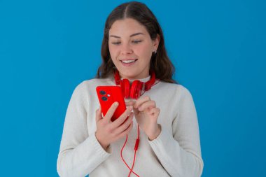 Boynuna kırmızı kulaklık takmış, akıllı telefonuna bakıp gülümseyen mutlu beyaz bir kadın. Mavi arka plan onun neşeli ve meşgul yüz ifadesini vurguluyor.. 