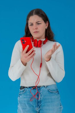 Beyaz kazaklı, elinde kulaklıkla kırmızı akıllı telefon tutan endişeli genç bir kadın, ekranı, alnı mavi arka planı izlerken şok ve stres içinde tepki veriyor.