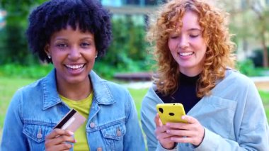  Neşeli Afrikalı Amerikalı ve Kafkasyalı arkadaşlar ellerinde renkli akıllı telefonlar ve bir kredi kartı ile bir şehir parkında duruyorlar. Nakitsiz kolaylığı kutluyorlar, alışveriş başarılarını paylaşıyorlar ve dijital bir yaşam tarzları var.