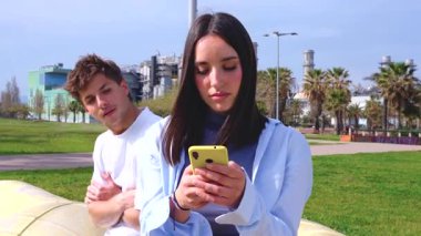  Genç çift sahil kenarındaki bankta oturuyor, kadın sarı akıllı telefona odaklanıyor, dikkatle kayıyor, erkek arkadaşı ise görmezden gelindiği için üzgün, dijital dikkat dağınıklığı ve ilişki gerilimini resmediyor..