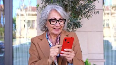 Gri saçlı ve gözlüklü neşeli yaşlı kadın modern bir binanın dışında kırmızı bir akıllı telefon kullanıyor. Ceket ve desenli eşarp giyiyor. Bağlantının keyfini çıkarıyor. 