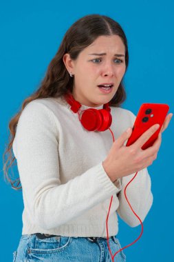 Beyaz kazaklı, elinde kulaklıkla kırmızı akıllı telefon tutan endişeli genç bir kadın, ekranı, alnı mavi arka planı izlerken şok ve stres içinde tepki veriyor.