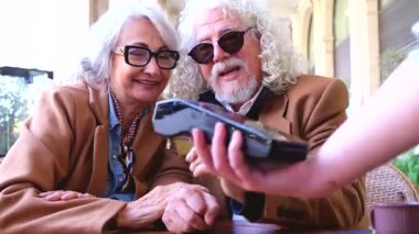 Gülümseyen son sınıf çifti açık hava kafesinde kredi kartı ödemesi yapıyor. Şık palto ve güneş gözlüğü takıyorlar, güneşli, günlük ve kentsel bir ortamda rahat bir anın tadını çıkarıyorlar.. 