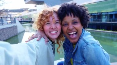Heyecanlı Afrikalı Amerikalı ve Kafkasyalı arkadaşlar omuza sarılıyor, modern şehir kanalının yanında cam mimarili parlak bir selfie çekmek için kollarını uzatıyor, arkadaşlığı, seyahati ve paylaşılan anıları kutluyorlar.. 