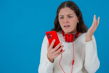Beyaz kazaklı, elinde kulaklıkla kırmızı akıllı telefon tutan endişeli genç bir kadın, ekranda, alnında, mavi arka planda izlerken şok ve stres içinde tepki veriyor. Boşluğu kopyala 